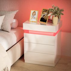 a white nightstand sitting next to a bed with pink walls and pillows on top of it