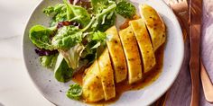 a white plate topped with sliced chicken next to a green salad and fork on top of a table