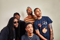 four men are posing for the camera in front of a white wall and one man has dreadlocks on his head