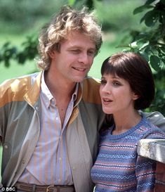 a man and woman standing next to each other in front of a tree with leaves