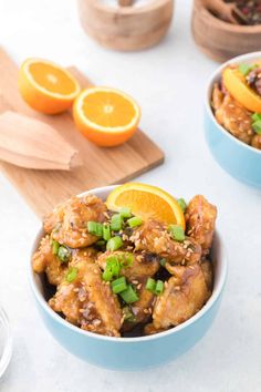 two bowls filled with orange chicken and garnished with scallions next to sliced oranges