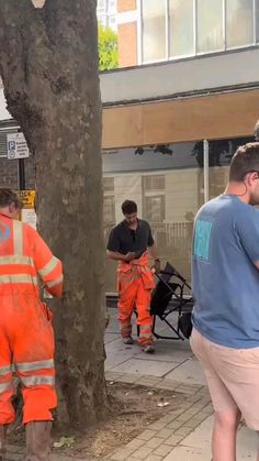 men in orange safety suits standing next to a tree on the sidewalk while another man looks at his cell phone