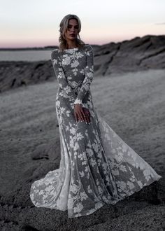 a woman in a long dress standing on the beach at sunset with her hands behind her back