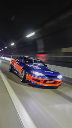 an orange and blue car is driving down the road at night time with its lights on