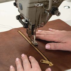 two hands using a sewing machine to sew on a piece of brown leather with gold zippers