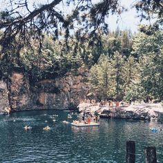 several people are swimming in the water near some rocks and trees, while others float on small rafts