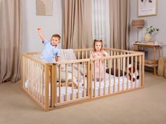 two children standing in a wooden crib with curtains on the windowsill and one child raising his hand