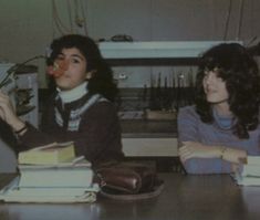 two young women sitting at a table with books and papers in front of their faces