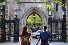 two people are walking down the sidewalk in front of an iron gate with cars behind them