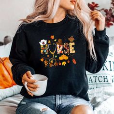 a woman sitting on a bed holding a coffee cup and wearing a black sweater with fall leaves