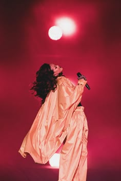 a woman in an orange outfit holding a microphone and singing into the air with her arms outstretched