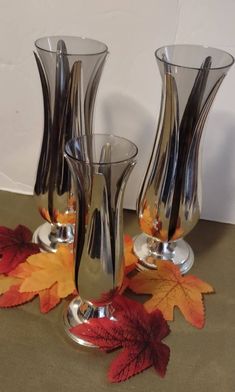 three silver vases sitting next to each other with leaves on the ground around them