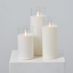 three white candles sitting on top of a white block with one lit candle in the middle