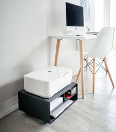 a desk with a computer and printer on it in front of a white chair next to a window