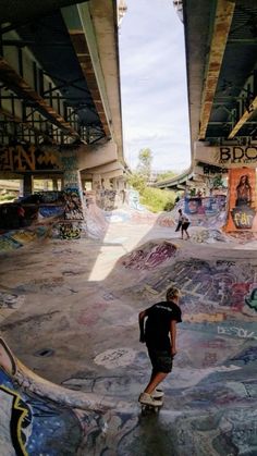 a skateboarder rides his board under an overpass in a skate park with graffiti on the walls