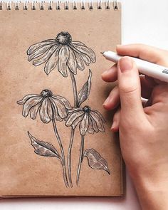 a hand is holding a pen and drawing flowers on a brown notebook with white paper