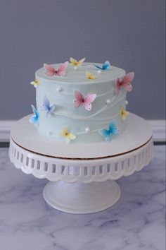 a white cake with blue frosting and colorful butterflies on the top is sitting on a pedestal