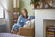 a woman sitting in a chair next to a fireplace with books on the mantle and potted plants