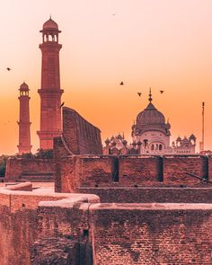 an old brick building with two towers and birds flying in the sky above it at sunset