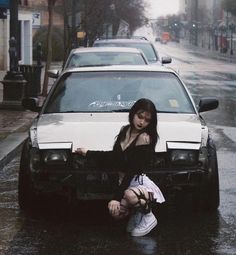 a woman sitting on the hood of a car in the middle of a rain soaked street