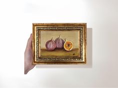 a person holding up a painting with an orange and two figs on the table