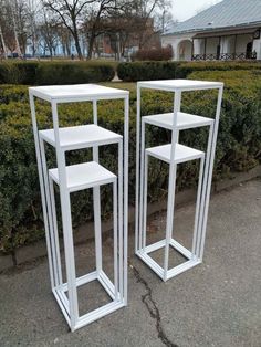 two white metal plant stands sitting next to each other in front of bushes and shrubbery