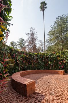 a circular brick bench sitting in the middle of a flowered area with trees and bushes behind it