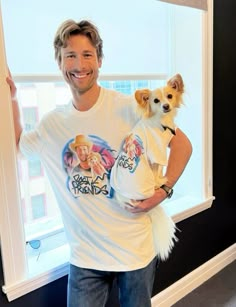 a man holding a small dog in his arms and smiling at the camera while standing next to a window