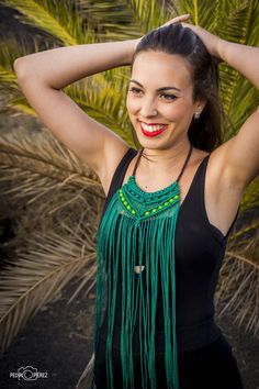 a woman in a black top and green fringe necklace smiles at the camera with her hands on her head