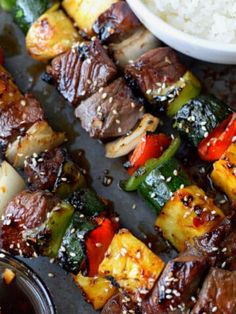 skewered steak and vegetables with sesame seeds on a black tray next to rice