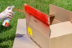 a person sprays red paint onto a cardboard box on the ground in front of some grass