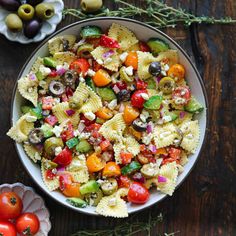 a bowl full of pasta salad with olives, tomatoes and other vegetables on the side