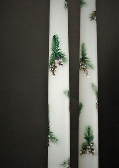 two white candles decorated with pine cones and evergreen needles