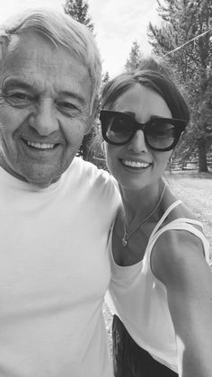 an older man and young woman posing for a photo in front of some pine trees