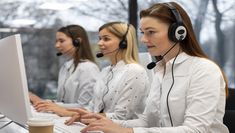 three women wearing headsets are sitting at a desk with computer monitors and laptops