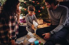 a man, woman and child sitting in front of a christmas tree with presents on the floor