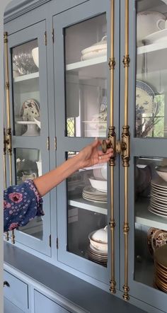 a woman reaching for plates in a blue china cabinet with gold trim and glass doors