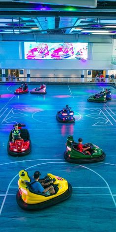 several people are riding bumper cars in an indoor area with lights on the ceiling and large screen behind them