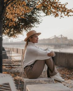 a woman sitting on top of a wooden bench next to a tree in the fall