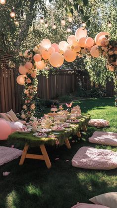 Outdoor fairy garden birthday party setup with a moss-covered table, soft pink cushions, whimsical balloon arch, string lights, and delicate floral decorations, creating a magical and enchanting atmosphere. Outdoor Fairy Birthday Party Ideas, Girls Garden Party Birthday, Spring Garden Birthday Party, Garden Party Theme Birthday, Intimate 1st Birthday Party Ideas, Wild And Wonderful Birthday Party, Garden Party First Birthday Girl, Fairy Decorations Party, Enchanted Fairy Garden Party Decoration