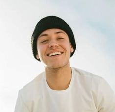 a young man wearing a beanie smiles at the camera while standing in front of a blue sky