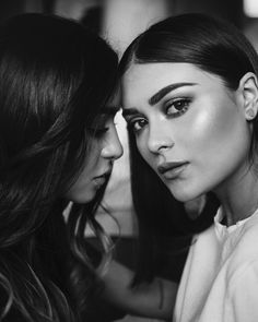 two beautiful young women sitting next to each other in front of a mirror, one looking at the camera