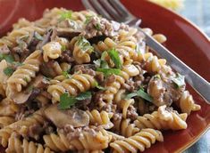 a red plate topped with pasta covered in meat and mushrooms next to a knife and fork