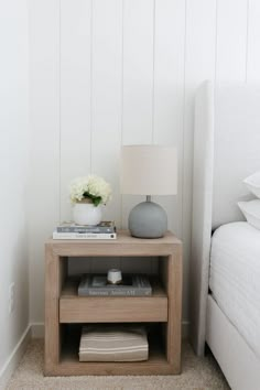 a nightstand with a lamp, books and flowers on it next to a white bed