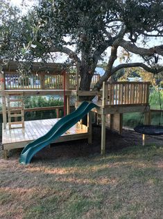 a wooden play set with a slide and swings underneath a large tree in a park