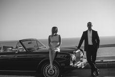 a man standing next to a woman in front of a black and white car near the ocean