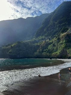 some people are walking on the beach by the water and mountains in the background,