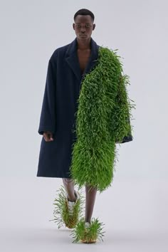 a man standing in front of a white wall wearing a coat and grass plant skirt