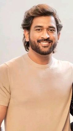 a man with long hair and beard standing in front of a white wall wearing a tan t - shirt