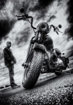 a black and white photo of a man standing next to a motorcycle on the road